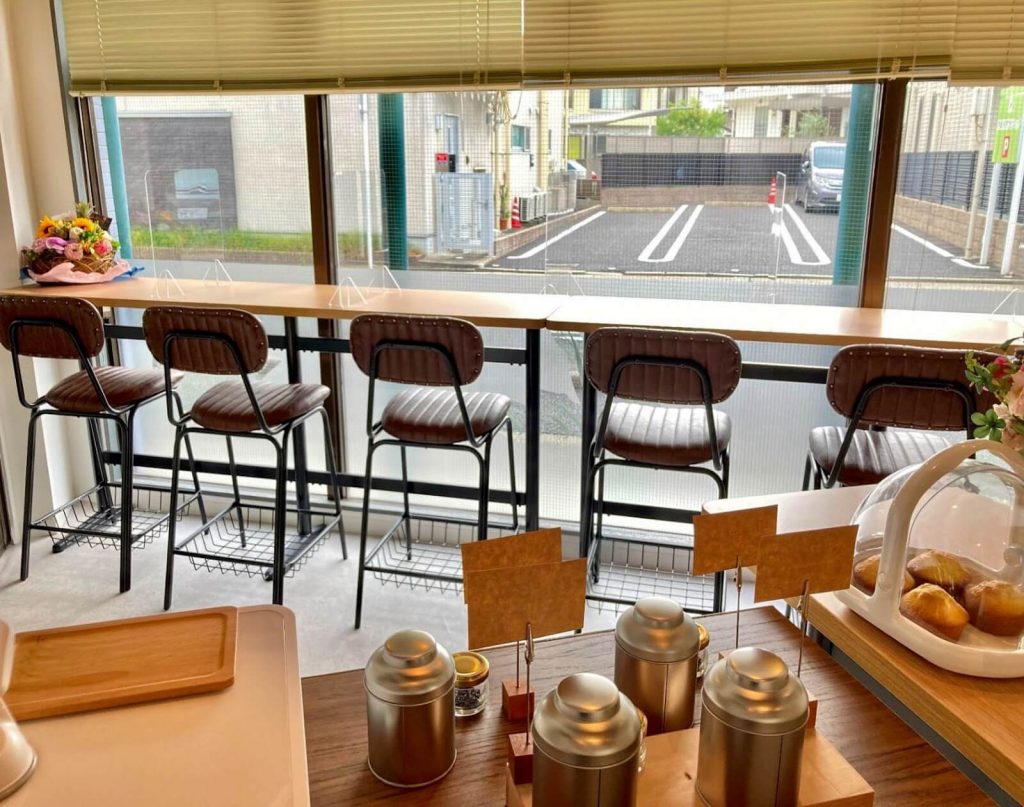 窓際にカウンター席が並び、手前には商品が陳列された棚があるカフェの店内。カウンター席には革張りのスツールが並び、窓の外には駐車場が見える。﻿