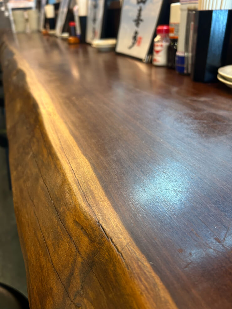 節や木目が特徴的な、艶のある木製のカウンター。奥には飲食店のような空間がぼやけて見える。