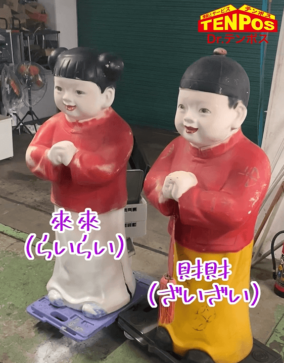 赤い上着を着た二体の招き人形。左の人形には「來來 (らいらい)」と書かれており、右の人形には「財財 (ざいざい)」と書かれています。